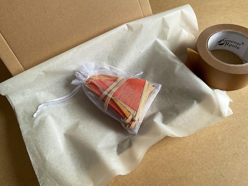 May include: A white tissue paper bag with a drawstring closure, filled with a stack of colorful paper pennants. The bag is sitting on a brown cardboard surface.
