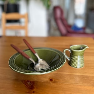 Puede incluir: Un tazón de cerámica verde con dos cucharas de plata y una jarra de cerámica verde sobre una mesa de madera.