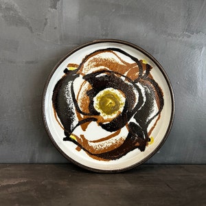 Può includere: Un piatto in ceramica con base bianca e bordo marrone. Il piatto presenta un design audace e astratto nei toni del marrone, nero e giallo. Il design ricorda un fiore o una spirale stilizzata. Un pezzo decorativo per la cucina o la sala da pranzo.