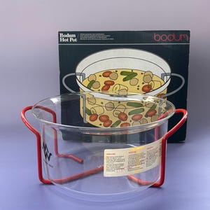 Pode incluir: Uma panela Bodum Hot Pot de vidro transparente com alças de metal vermelho e uma caixa correspondente no fundo. A caixa apresenta uma ilustração da panela cheia de comida. A panela é projetada para cozinhar e servir.