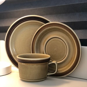 Puede incluir: Un juego de tazas y platillos de té de cerámica marrón y beige. La taza y el platillo tienen una franja marrón alrededor del borde.