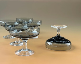 May include: A set of six gray-tinted coupe glasses. Five glasses are upright, showcasing their rounded bowls and stemmed bases. One glass is inverted, revealing its dark gray base. The glasses are arranged on a light orange surface.