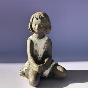 May include: A ceramic sculpture of a seated girl with short hair. The figure is primarily brown with white accents on the dress. The sculpture is kneeling with hands clasped, and the overall aesthetic is rustic.