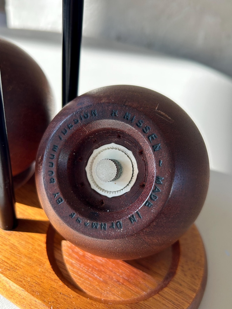 May include: A close-up of a wooden salt shaker with a white grinder. The bottom of the shaker is engraved with "BOOM DESIGN" and "MADE IN DENMARK". The shaker is part of a set on a wooden stand.