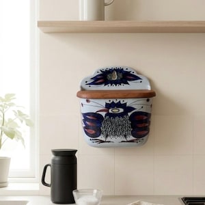 Puede incluir: Un recipiente de cerámica montado en la pared con una tapa de madera. El recipiente es de color azul claro con un diseño pintado a mano de un pájaro en tonos azules y rojos. El recipiente está montado en una pared de color crema. Una taza negra y un cuenco de cristal pequeño están en la encimera debajo.