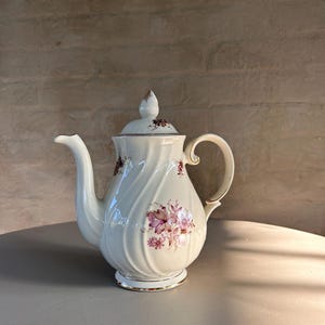 Puede incluir: Tetera de porcelana blanca con un diseño floral en tonos rosa y marrón. La tetera tiene un borde dorado alrededor del borde y un asa dorada.
