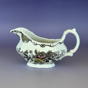 May include: A white ceramic gravy boat with a floral pattern in black, pink, and yellow. The gravy boat has a curved spout and a handle. The floral design is on the body of the boat. The rim is decorated with a black and gold design.