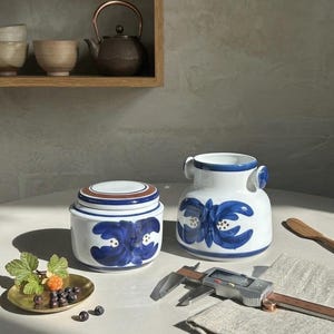 Op de afbeelding: Twee witte keramische containers met blauwe bloemmotieven. Eén is een pot met deksel, de andere een vaas met handvatten. Een klein messing schaaltje met bessen en een digitale schuifmaat staan ook op de tafel.