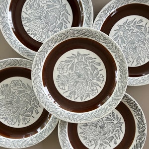 May include: Set of five white ceramic dinner plates with a brown and silver floral design. The plates have a raised rim and a scalloped edge.