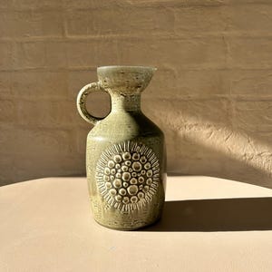 Op de afbeelding: Een groene keramische kan met een getextureerd oppervlak en een cirkelvormig ontwerp. De kan heeft een handvat en een smalle hals.