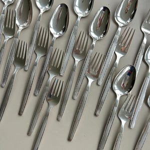 Può includere: Un set di posate d'argento, tra cui cucchiai e forchette, con un design testurizzato. Le posate sono disposte in uno schema sfalsato su una superficie grigio chiaro.