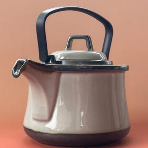 Pode incluir: Uma chaleira de cerâmica creme com base castanha escura e alça preta. A chaleira tem um bico e tampa prateados. A alça arqueia-se sobre a parte superior. O fundo é de cor pêssego lisa. Ideal para chá.