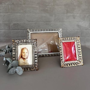 May include: Three ornate gold picture frames with decorative edges. The frame on the left has a photo of a young girl with blonde hair. The middle frame is empty. The frame on the right has a red background.