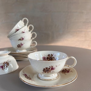 Puede incluir: Un juego de tazas de té y platillos vintage con un patrón floral en tonos de rosa y marrón. Las tazas y los platillos son blancos con un borde dorado.