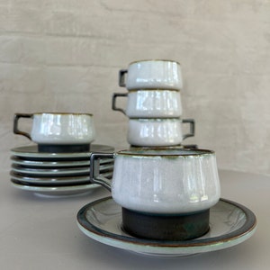 May include: A set of white ceramic teacups and saucers. The cups have a square handle and a dark rim. The saucers have a dark base and a light rim. The cups are stacked in a group of four, with additional cups and saucers visible.
