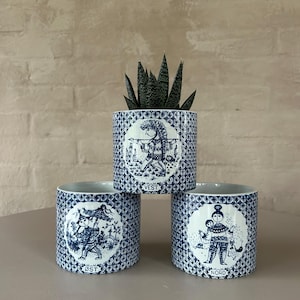 May include: Three blue and white ceramic planters with a folk art design. The planters are stacked with a succulent plant in the top planter. The planters are labeled "Vest", "Ost", and "Nord".