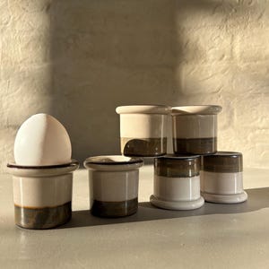 Può includere: Una collezione di portauova in ceramica in una palette di colori neutri. I portauova sono bianchi con una fascia marrone alla base. Un portauovo contiene un uovo bianco. I portauova sono disposti su una superficie grigio chiaro.