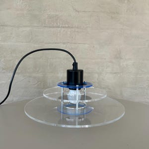 Könnte beinhalten: Eine moderne Pendelleuchte mit einem schwarzen Kabel und einem transparenten Acrylschirm. Der Schirm hat drei Ebenen und einen blauen Akzentringe. Die Lampe ist eingeschaltet und eine Glühbirne ist sichtbar.