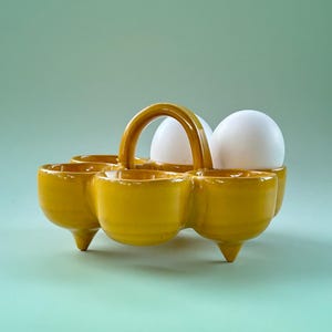 May include: A yellow ceramic egg holder with a handle, holding two white eggs. The holder has six egg cups and three small feet. The background is a solid light green colour.