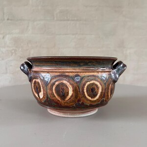 May include: A brown ceramic bowl with two handles and a white and brown geometric pattern. The bowl has a slightly rounded bottom and a smooth, glazed surface.