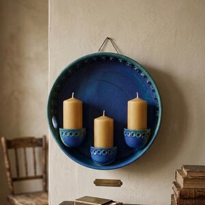 Puede incluir: Un candelabro de pared circular de cerámica azul con un esmalte cobalto profundo y un borde festoneado decorativo. Tres copas semicirculares integradas sostienen velas de cera cilíndricas de color tostado claro. Incluye un lazo de metal para colgar.