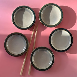 Può includere: Quattro ciotole rotonde in ceramica con centro grigio chiaro e bordo verde scuro, disposte in cerchio. Due bacchette di legno chiaro sono posizionate in diagonale tra le ciotole, puntando verso l'angolo in alto a sinistra.