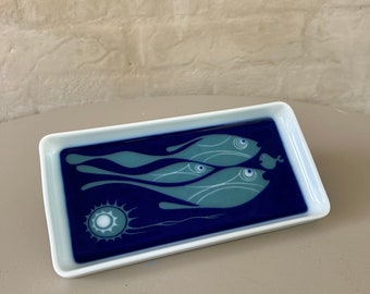 May include: A white rectangular ceramic dish with a blue and green design of fish swimming in the water. The fish have white eyes and are outlined in white. The water is a dark blue color and there is a white sun in the bottom left corner.