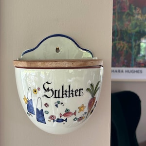 May include: A white ceramic wall-mounted container with a wooden lid. The container is decorated with colorful illustrations of fish, stars, and a carrot. The word "Sukker" is written in black script.