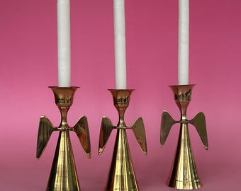 May include: Three polished brass candle holders shaped like stylized angels with conical bases and outstretched wings. Each holder supports a single, slender, white taper candle. The metallic finish is reflective and smooth.