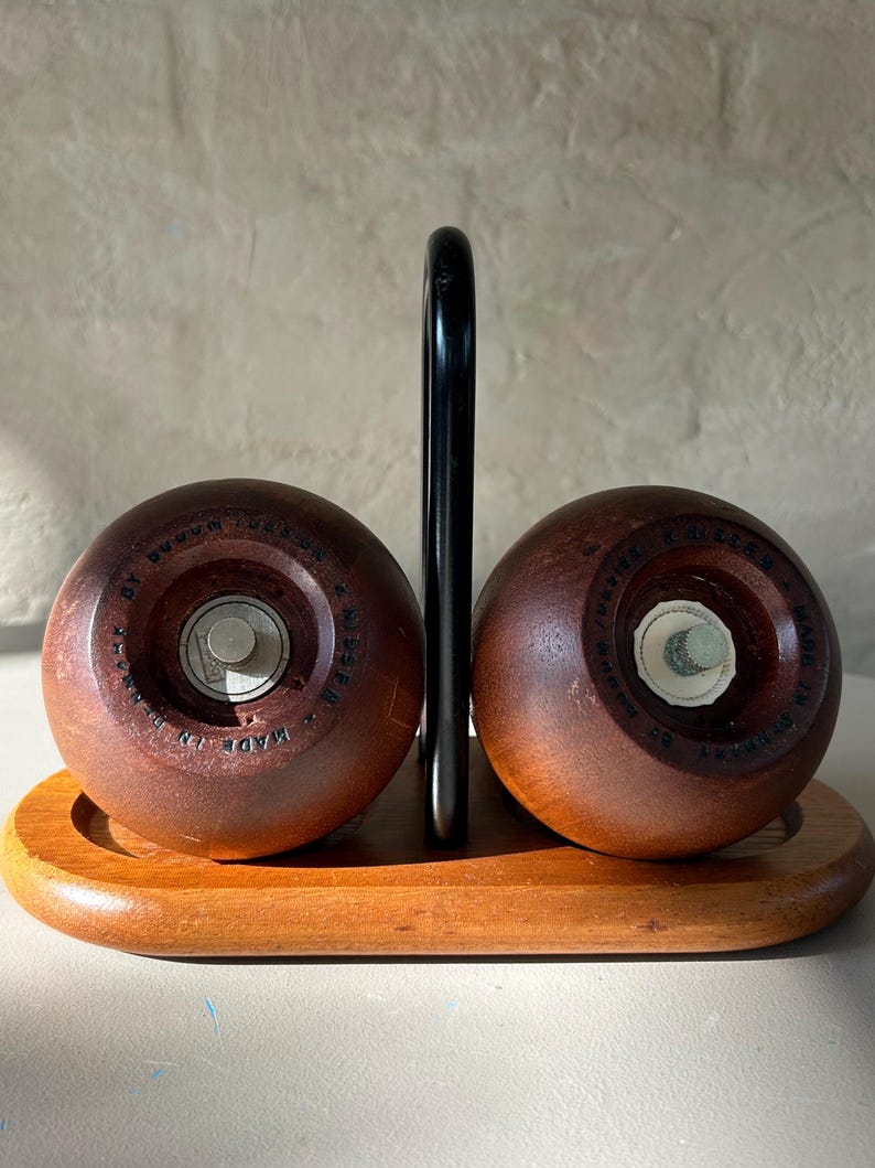 May include: A vintage salt and pepper shaker set. The set includes two round, dark brown wooden shakers with metal mechanisms, resting on a wooden tray. A black metal handle is between the shakers. The words "MADE IN DENMARK" are visible on the shakers.