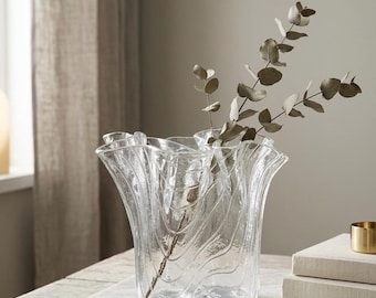 May include: A clear glass vase with a wavy, rippled rim and a textured, organic surface pattern. The vase features a wide, flared opening and a rounded base, containing a single stem with small, round, muted green leaves extending upward.