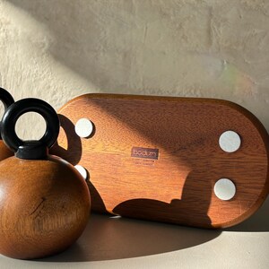May include: A wooden bottle stopper with a black handle and a round, brown body. A wooden serving tray with white pads is also visible. The tray has the brand name "bodum" printed on it.