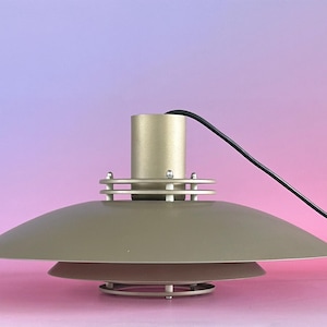 Puede incluir: Una lámpara colgante de color beige mate con una estructura de pantalla de metal ancha, poco profunda y de varios niveles. La lámpara incluye una carcasa cilíndrica central, un cable de alimentación negro y dos anillos metálicos.