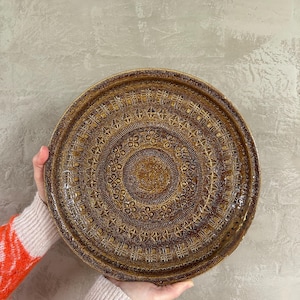 May include: A large, round, brown ceramic serving tray with a decorative, embossed pattern. The pattern features concentric circles of geometric designs.