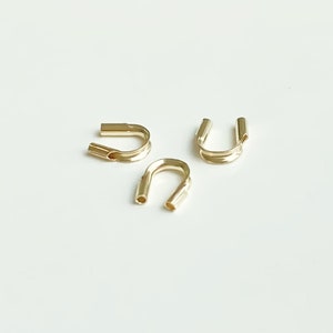 May include: Three gold-coloured metal crimp beads, each shaped like a U, are arranged on a white surface.