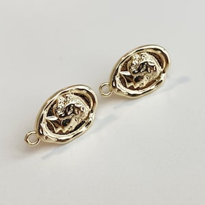 Peut inclure: Deux breloques ovales dorées avec un motif en relief d'un visage. Les breloques ont une boucle sur un côté pour les attacher à des bijoux.