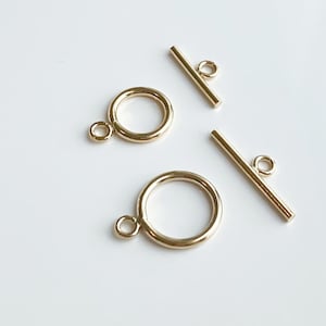 Op de afbeelding: Twee goudkleurige metalen schuifgespen met een ronde ring aan het ene uiteinde en een rechte staaf aan het andere uiteinde. De gespen worden vaak gebruikt voor het maken van sieraden.