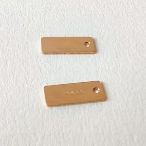 May include: Two gold-coloured metal rectangular tags with a hole at the top. The tag on the bottom has the text "100% Made" on it.