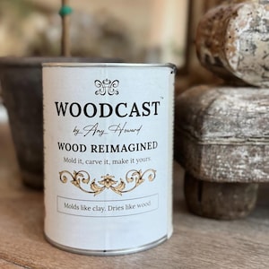 May include: A cylindrical can of "WOODCAST by Any Howard" with the text "WOOD REIMAGINED". The can has a white label with black text and a gold floral design. The bottom of the label reads "Molds like clay. Dries like wood."