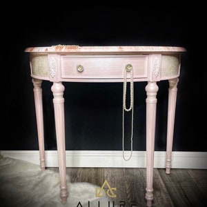 May include: A pink painted oval console table with a single drawer and ornate legs. The table top is marble with a pink and white pattern. A pearl necklace is draped over the table.