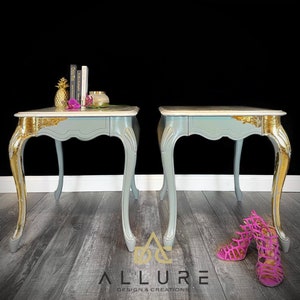 May include: Two matching light blue end tables with gold leaf accents and ornate legs. The tables have a white marble top and are sitting on a wood floor.  There are two pink high-heeled shoes in the foreground.