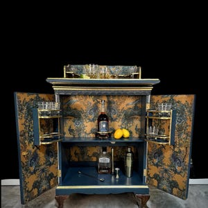 May include: A navy blue and gold bar cabinet with open doors, revealing a patterned interior. The cabinet features shelves with glassware, bottles of liquor, and bar tools. The doors and interior are decorated with a gold and blue floral pattern.