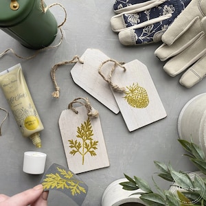 May include: Two white wooden tags with gold metallic paint, one with a strawberry design and the other with a leaf design. A bottle of gold metallic paint, a roll of twine, and a pair of gardening gloves are also in the image.