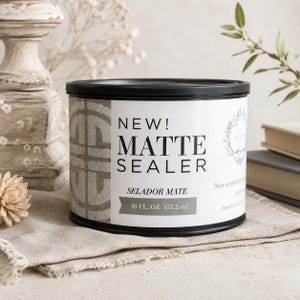 May include: A cylindrical container of matte sealer with a black lid and base. The white label features the text NEW! MATTE SEALER, SELADOR MATE, and 16 FL. OZ. 473.2 ml. The design includes a grey geometric pattern on the left and black typography.