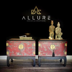 May include: Two red wooden nightstands with black legs and gold accents. Each nightstand has two doors with gold handles and a tassel. The nightstands are decorated with a distressed paint finish. Two terracotta warrior figurines are on top of the nightstands.