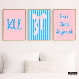 Op de afbeelding: Drie ingelijste prints met een lichte houten afwerking. Eén roze print heeft de letters "KU" in blauw. De middelste print heeft een roze strik op een blauw gestreepte achtergrond. De derde roze print zegt "Rock Chalk Jayhawk" in blauw.