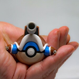 May include: A spherical toy figure featuring a brown lower half and a white upper section with a blue band. The object has a central circular button, two small blue side protrusions, and a prominent gray cylindrical nozzle extending from the top.