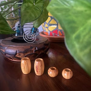 Puede incluir: Cuatro pequeñas cuentas de madera de color claro con un patrón de rayas se encuentran sobre una superficie de madera oscura. Las cuentas son cilíndricas y tienen una veta de madera natural. Un cuenco de madera tallada y un objeto metálico en espiral están en el fondo.