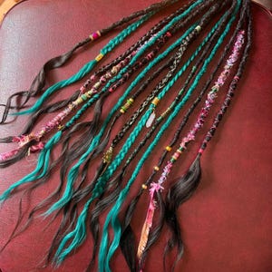 May include: A set of dark brown synthetic dreadlocks featuring teal and pink accents. The strands are decorated with various metallic beads, small charms, and textured yarn wraps. Some strands are braided, while others are loose and wavy.
