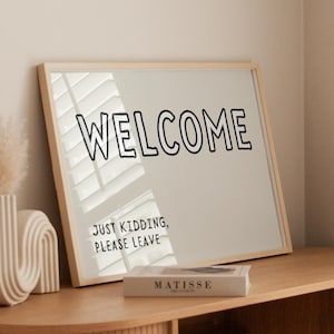 Op de afbeelding: Een ingelijst afdruk met de tekst "WELCOME" in grote, vetgedrukte letters. Onder de tekst staat de zin "JUST KIDDING, PLEASE LEAVE" in kleinere letters. De afdruk hangt aan een muur boven een houten bureau met een boek met de titel "MATISSE" erop.